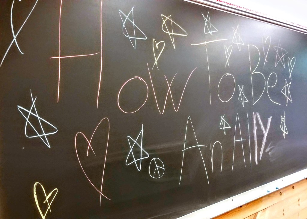 A blackboard that reads "How to Be an Ally" decorated with stars, hearts, and peace signs.