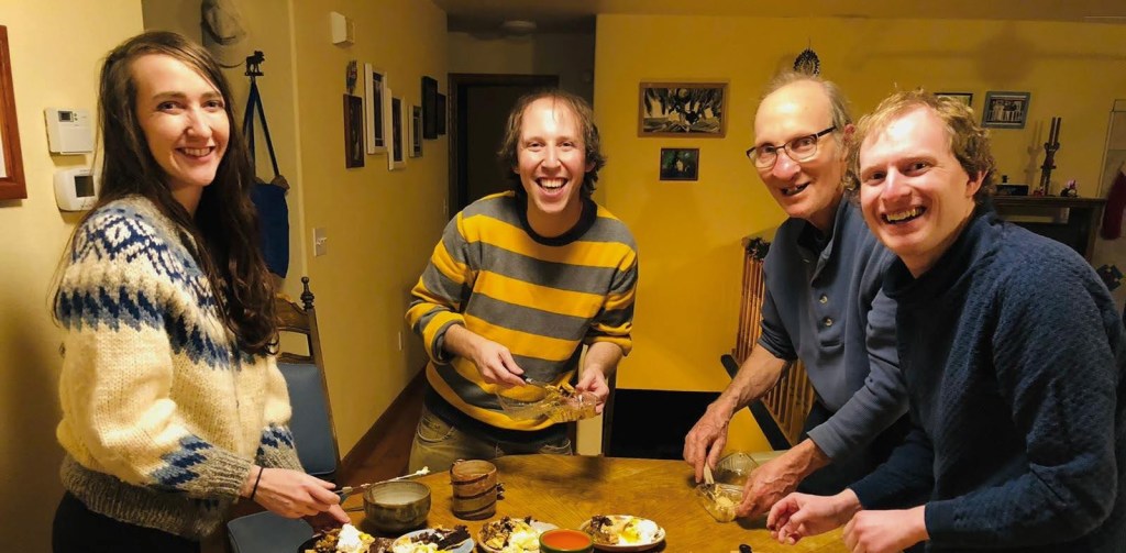 A photograph of four laughing people standing around a kitchen table.