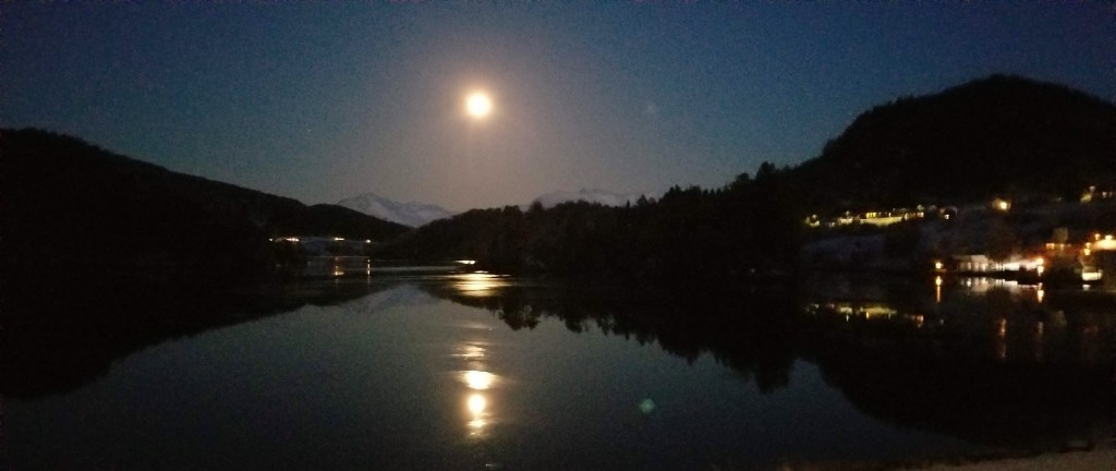 The moon reflected on water over snow-covered mountains.
