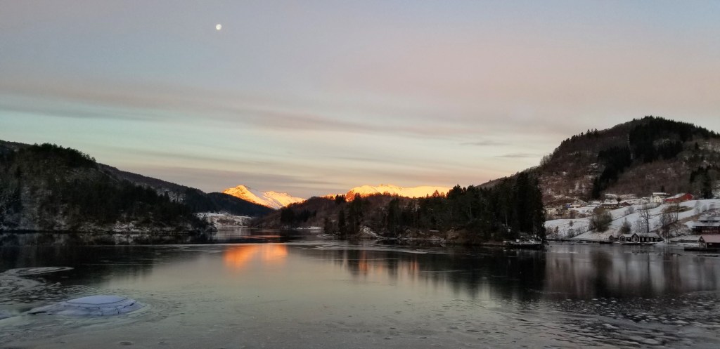 The moon above snow-covered mountains that are lit up red by the sun.