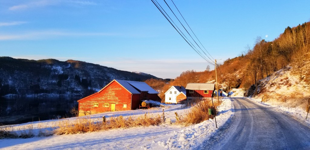 Sunlight on a barn, with a narrow road, electric lines, and the moon.