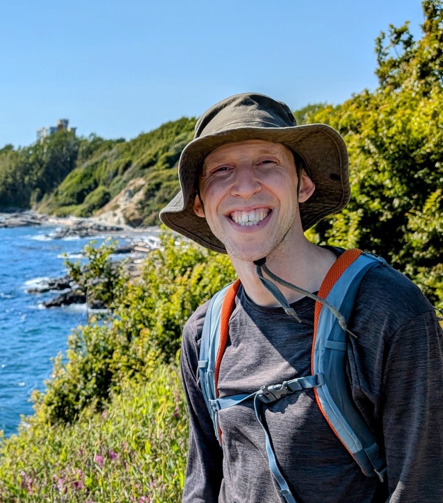 Jimmy Kindree standing in front of a green hillside and coast.